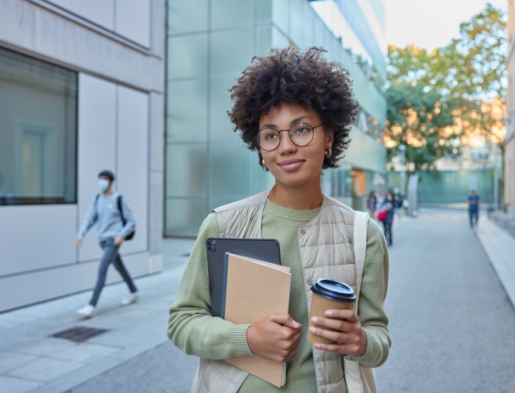 Beautiful curly female student holds notebook digital tablet and takeaway coffee focused into distance poses in urban setting wears casual clothes poses at busy street being on way to university